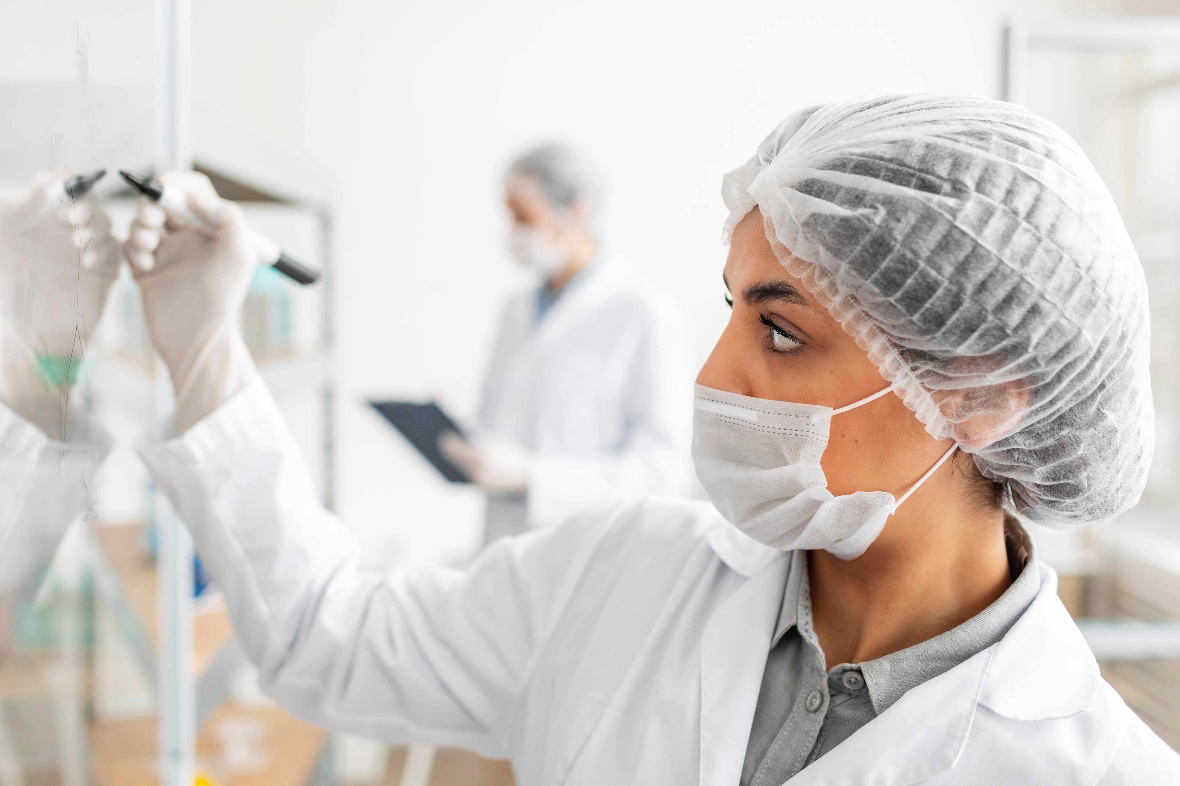 A technician in cleanroom clothing taking notes beside a stainless steel production vessel